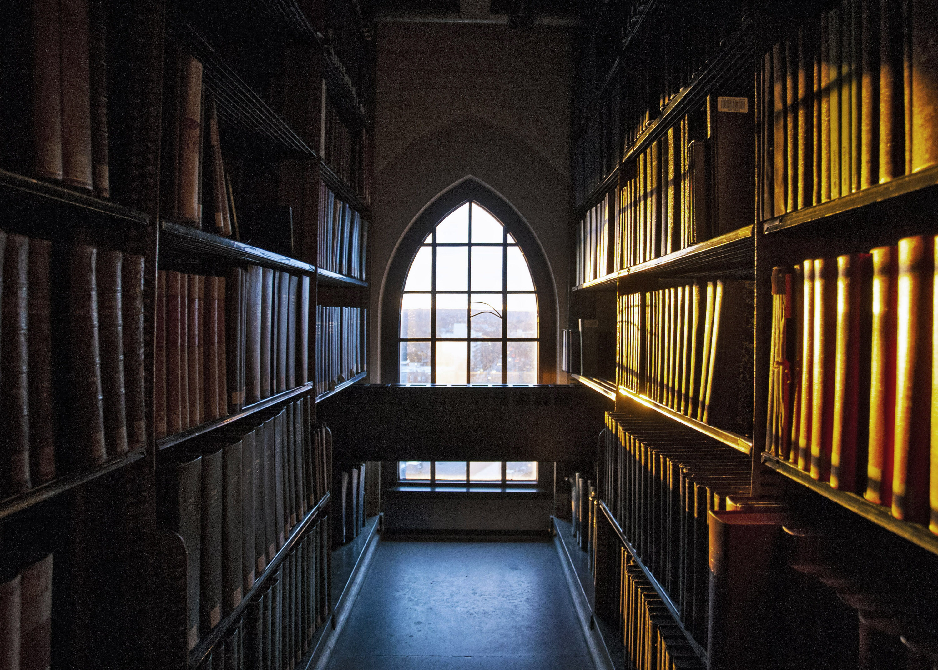 Dark aisle between shelves of books in a library looking towards an arched window